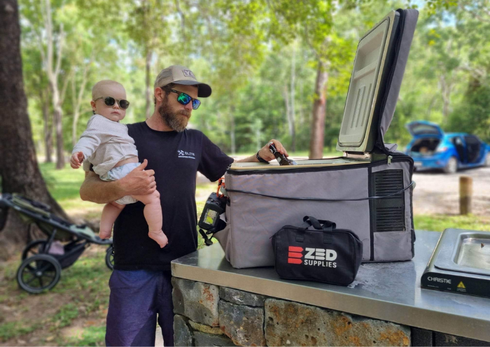 Dad holding his child at a park, a 12V camping fridge open on the BBQ bench kept cold by a ZED battery adapter